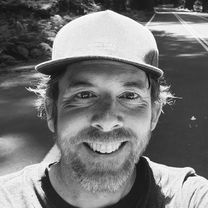 Colin, smiling, in a baseball cap on a tree-lined road. Black and white.